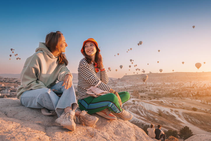 two omen sitting in a mountain looking at each other withhot air balloons in the sky in the background