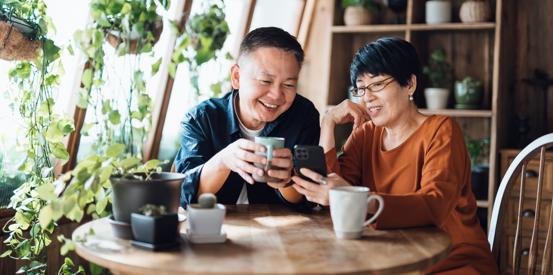 Older couple sitting at a table smiling down at a phone. 